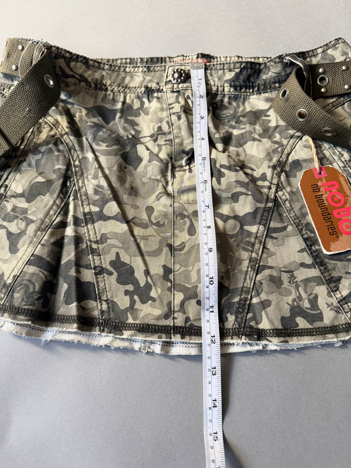 Photo of y2k camo mini skirt with original tags still attached and measuring tape showing 12 inches length.