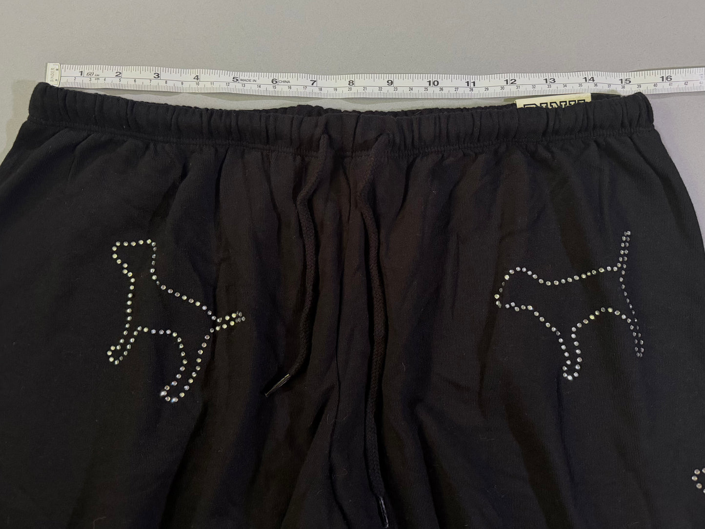 Black drawstring sweatpants with Victoria’s Secret pink rhinestone dogs and measuring tape showing waist measurement 