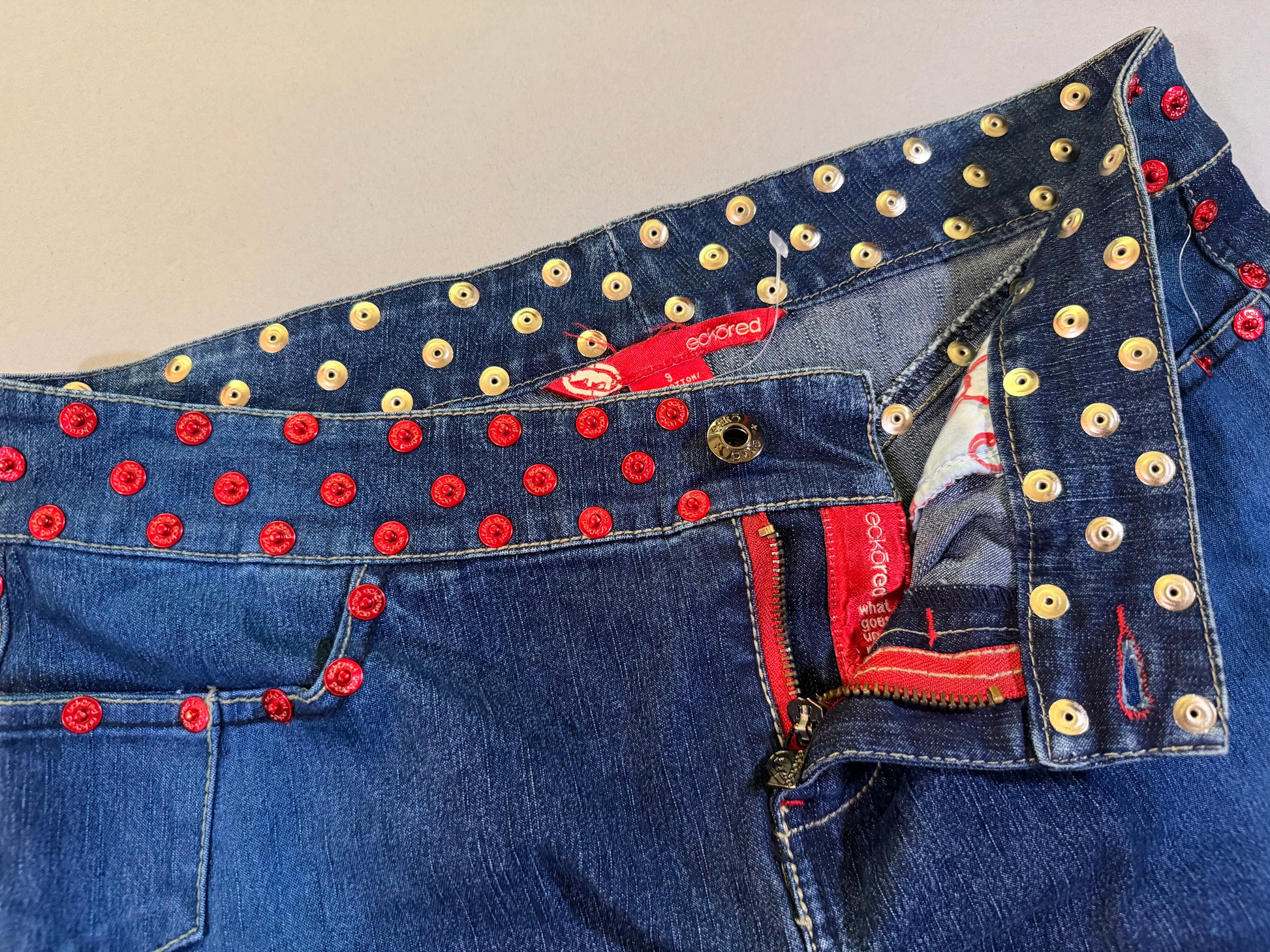 Close up of vintage denim skirt with red rivets showing open button and zipper fly 