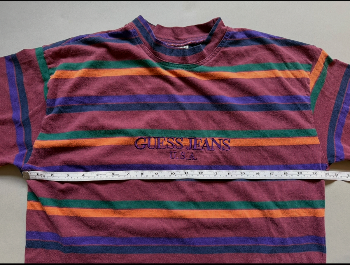 A vintage Guess Jeans short-sleeve t-shirt with horizontal stripes in purple, orange, yellow, and green, featuring the 'GUESS JEANS' logo across the chest. A measuring tape is placed horizontally across the chest to show the size.