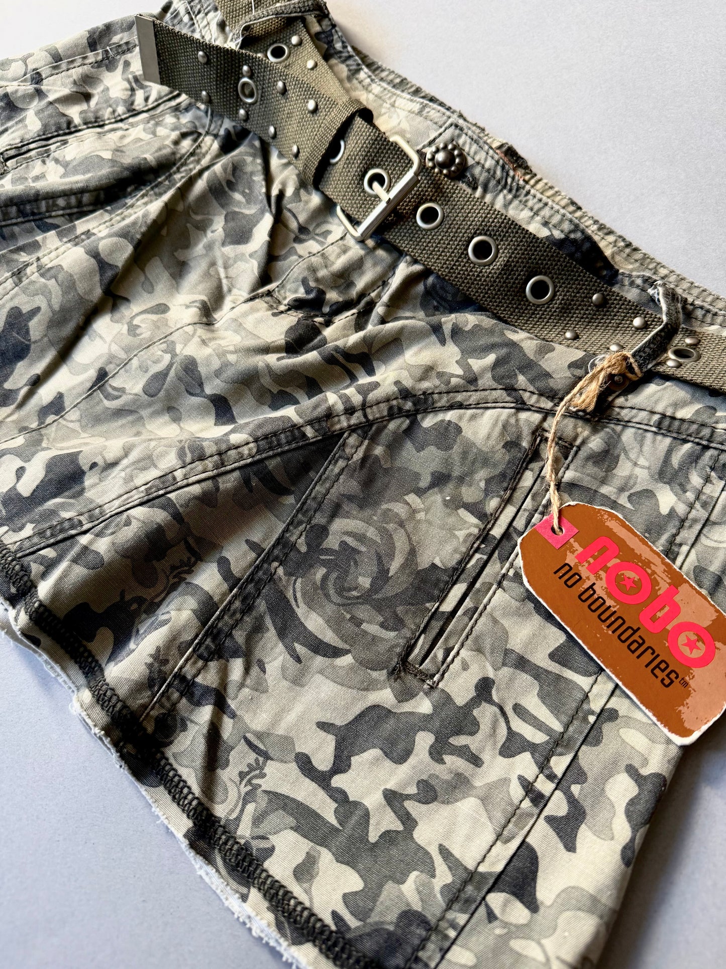 Angled Photo of y2k camo mini skirt with original tags still attached, showing more detail of matching studded grommet belt. 
