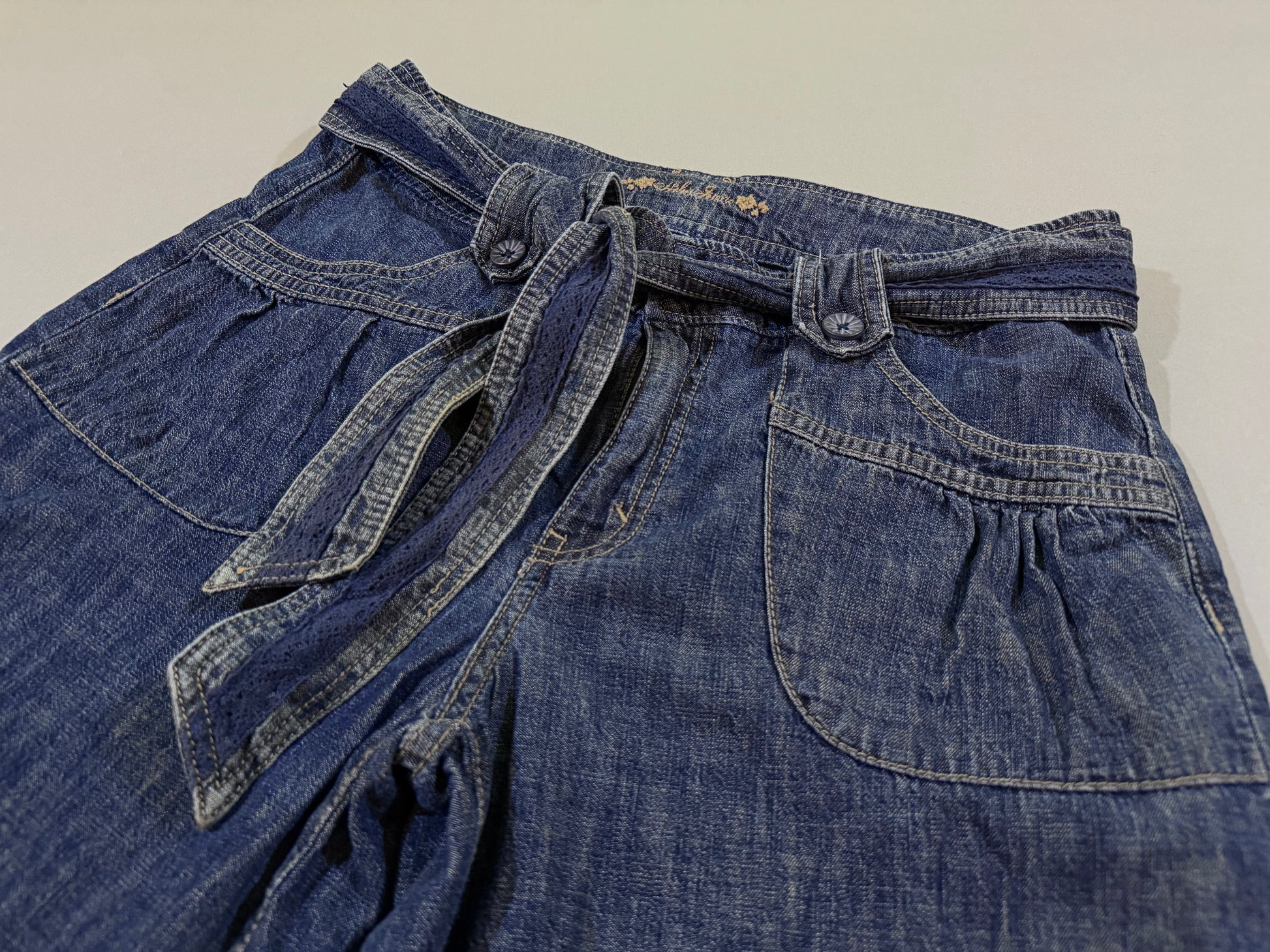 Close-up of the paperbag-style waist, removable denim belt, decorative buttons, and front patch pockets.