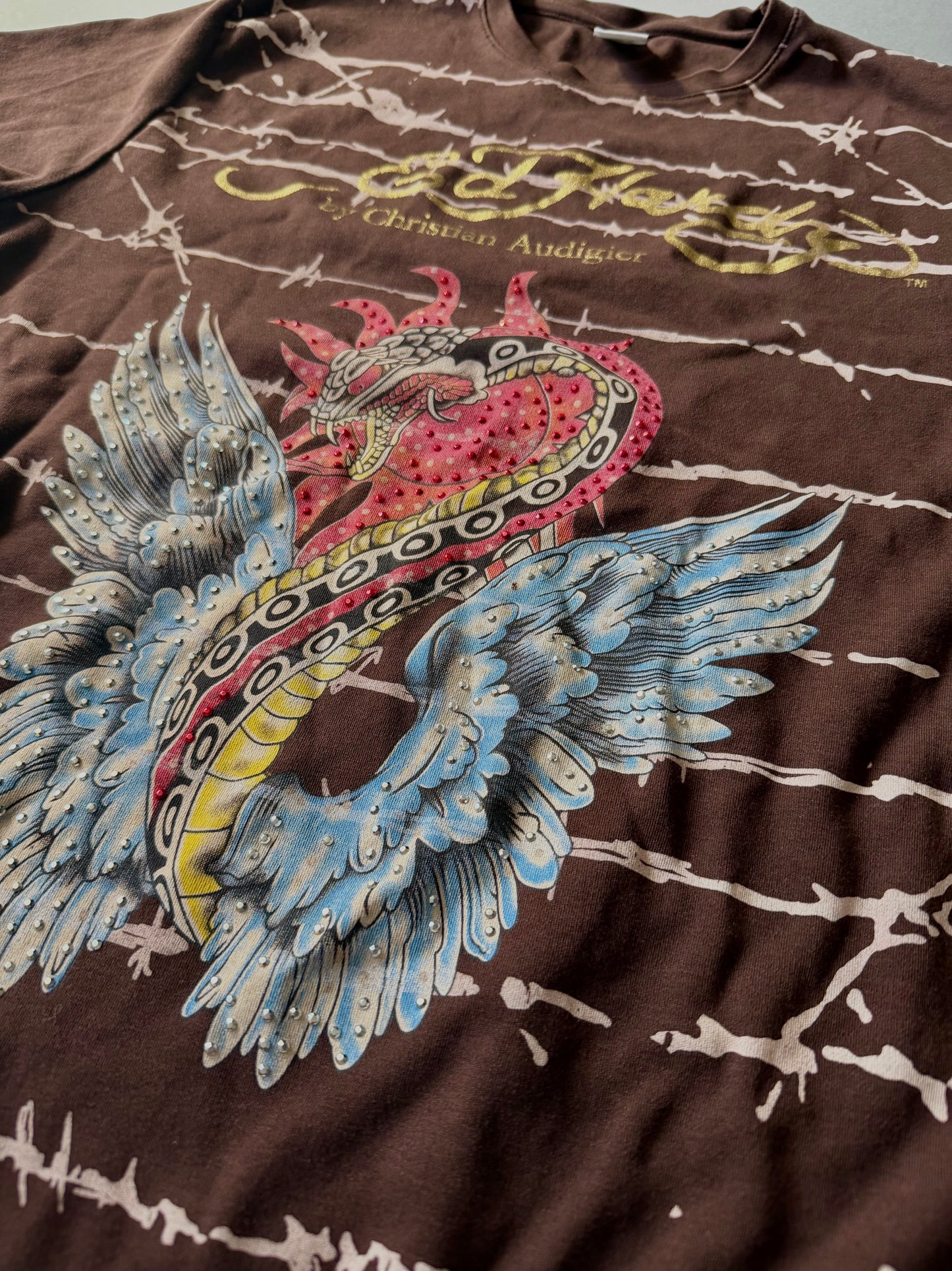 Angled photo of brown Ed hardy t-shirt with all over barbed wire print, close-up to the winged snake graphic with rhinestones throughout. 