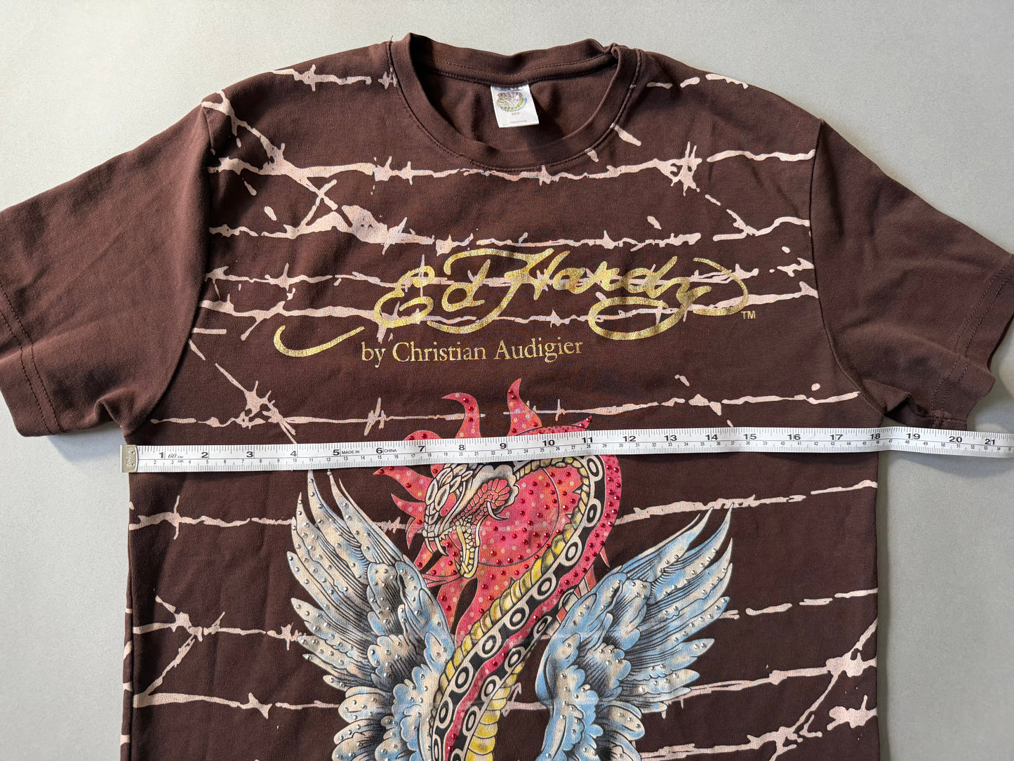 Photo of brown Ed hardy t-shirt with all over barbed wire print and winged snake graphic with rhinestones throughout. Measuring tape showing 18 inches pit to pit. 
