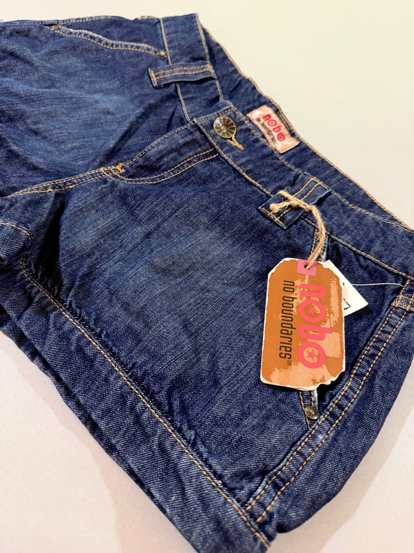 Angled close-up of Nobo Y2K denim shorts highlighting original hang tag, dark wash denim, and contrast stitching.