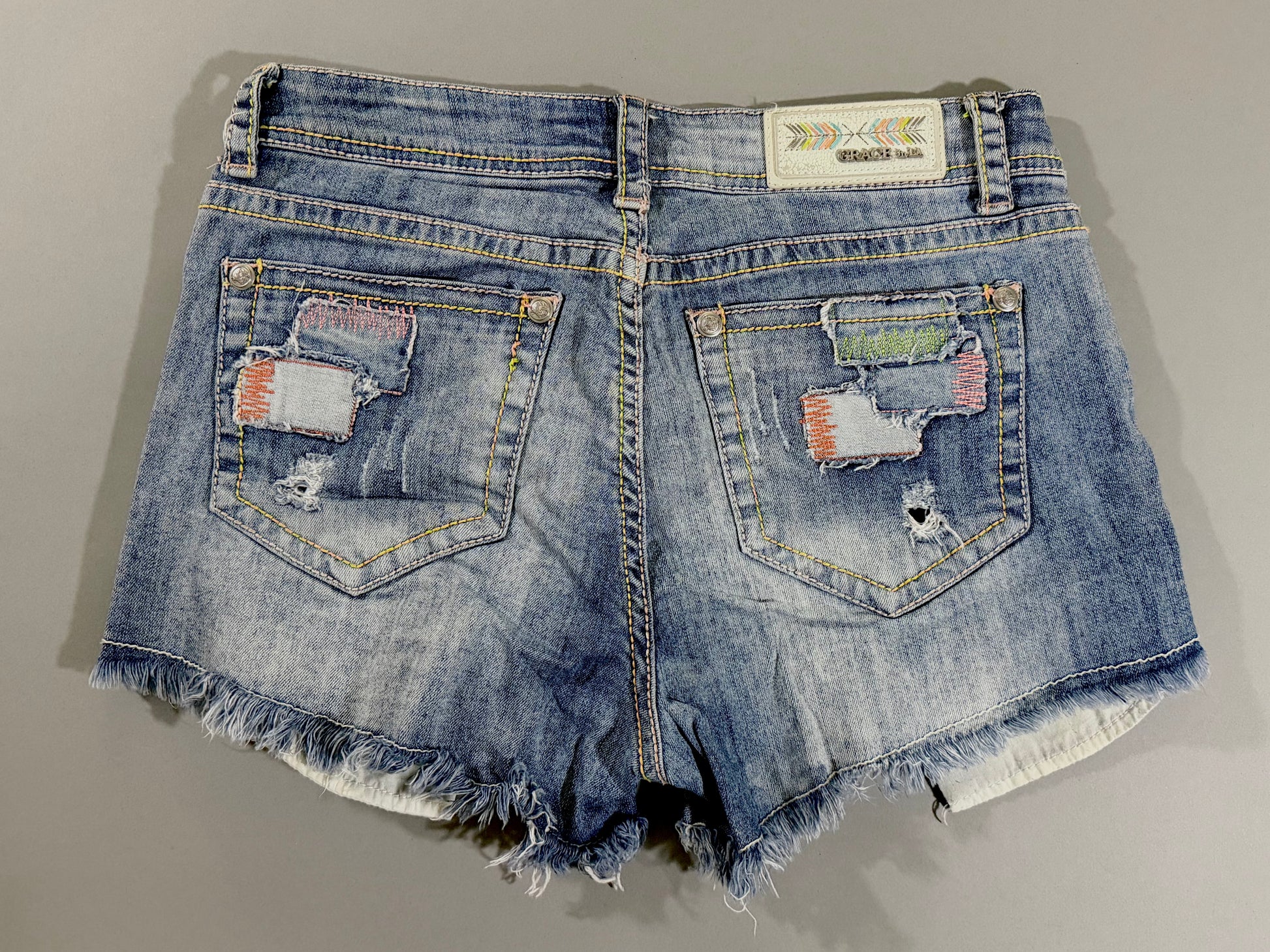 Back view of Grace in LA denim shorts with distressed back pockets, patchwork embellishments, holographic arrow logo patch, and raw frayed hems.