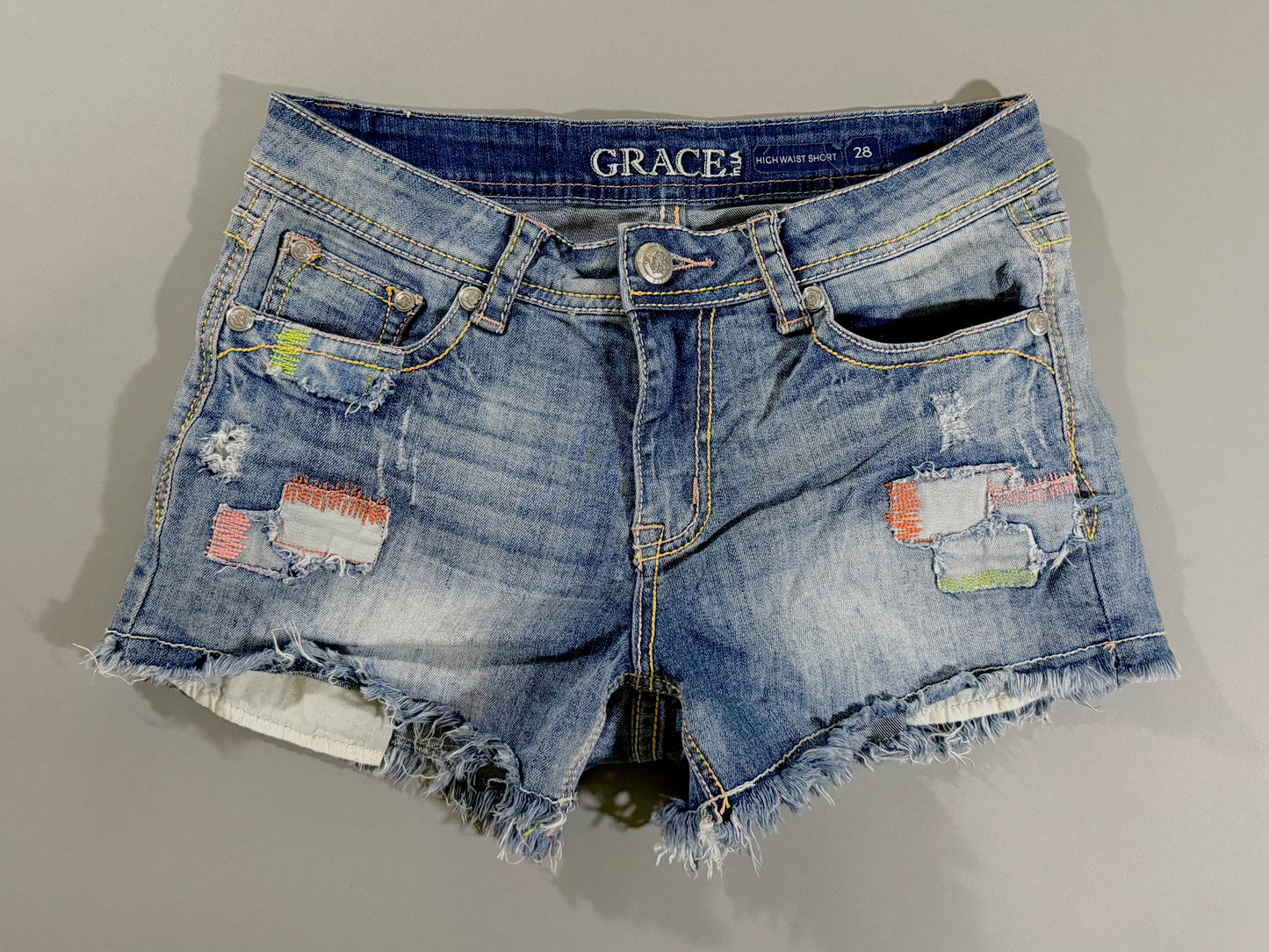 Grace in LA high rise distressed denim shorts in medium wash with colorful patchwork accents, frayed raw hem, and size 28 waistband.