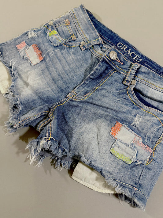 Angled front view of Grace in LA high rise jean shorts featuring distressed details, pastel patchwork, and colorful contrast stitching.