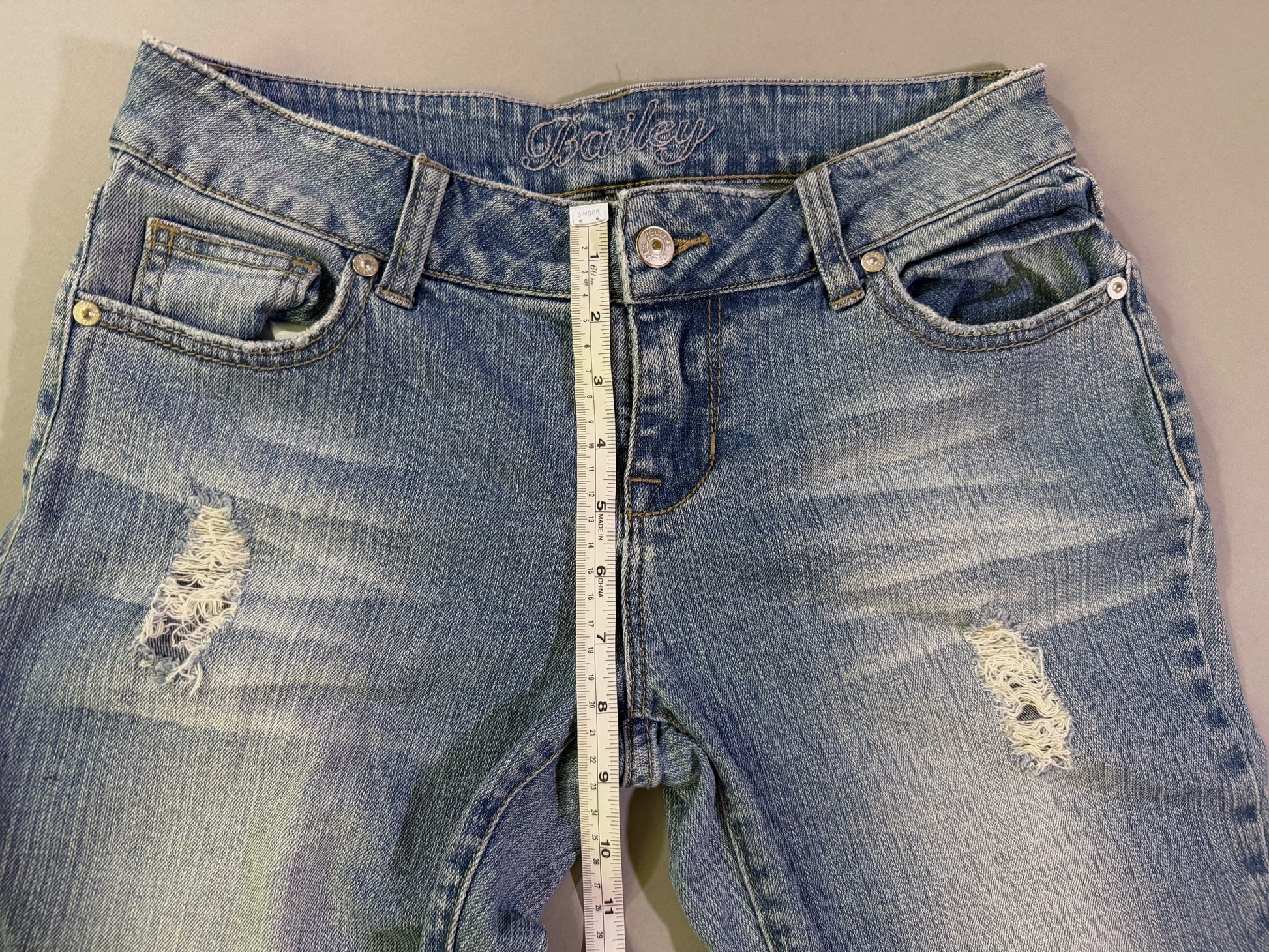Close up of waist of Delia’s bootcut blue jeans showing Bailey style name, distressing detail, and button/zip fly