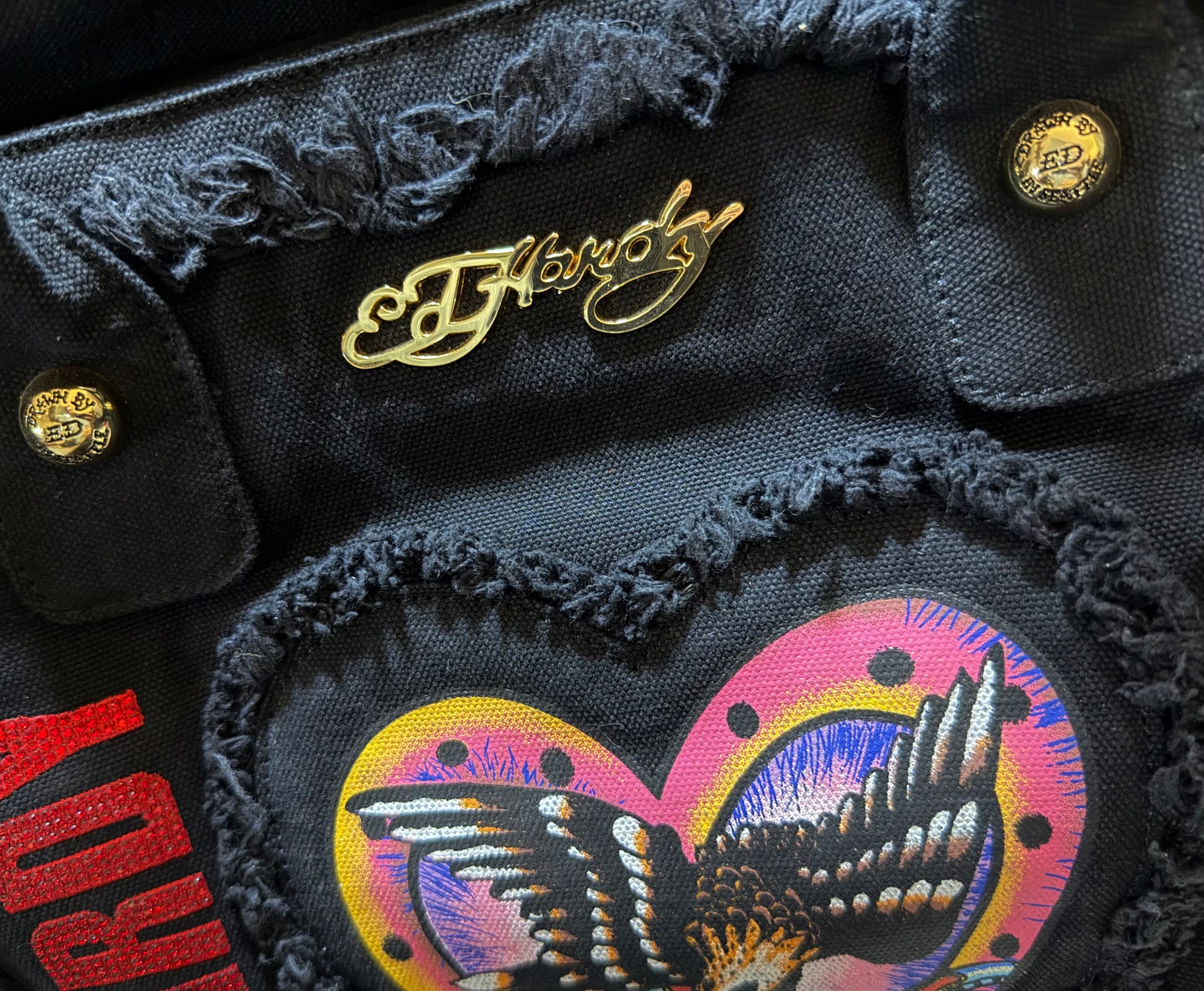 Close-up of gold Ed Hardy script hardware, frayed trim and colorful eagle tattoo artwork on the front panel.